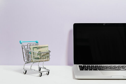 A Macbook Beside a Miniature Shopping Cart and Clipped Money